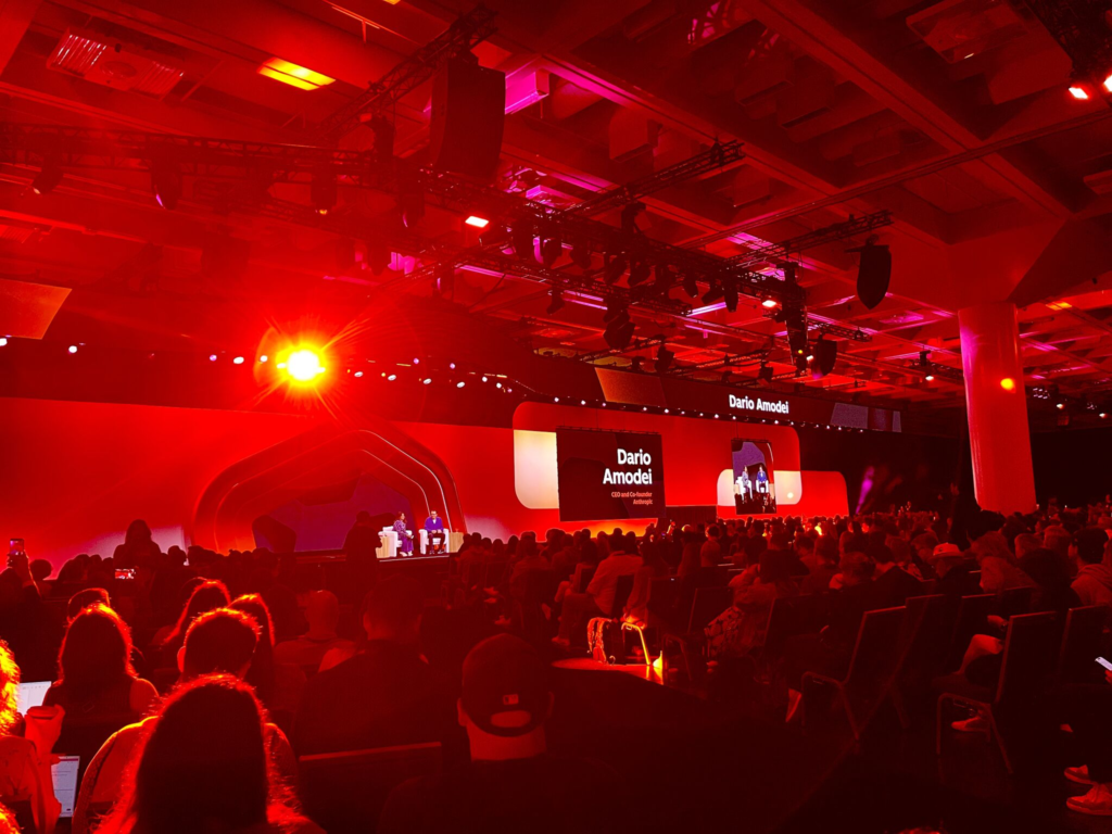 A packed auditorium bathed in red light, watching a speaker on the main stage