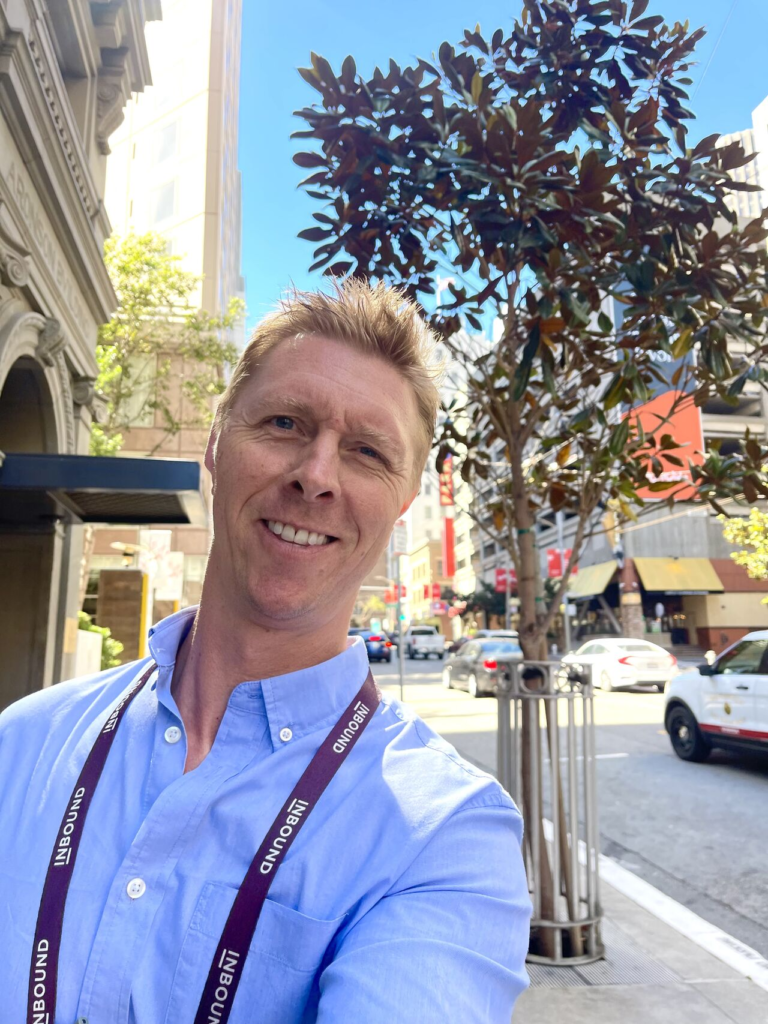 A smiling man wearing a conference lanyard takes a selfie on a city sidewalk