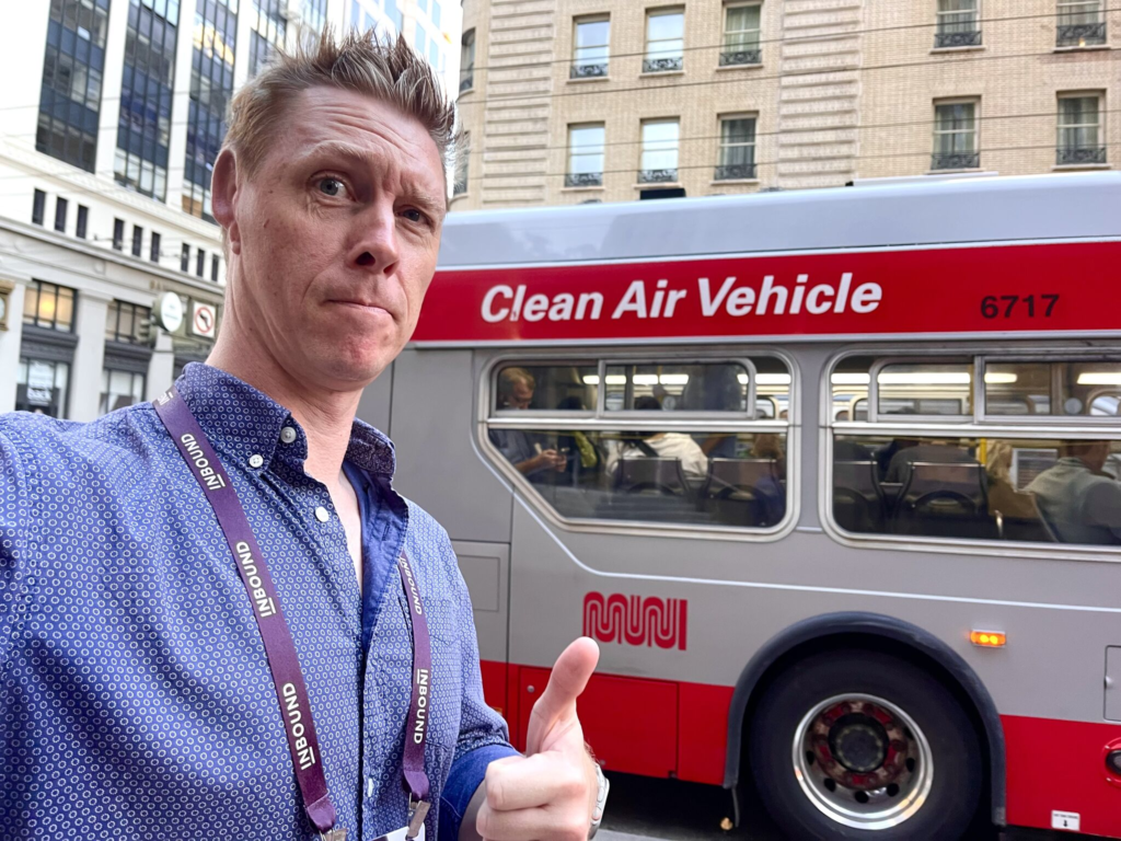 A man in a conference lanyard gives a thumbs-up in front of a red-and-white Clean Air Vehicle bus