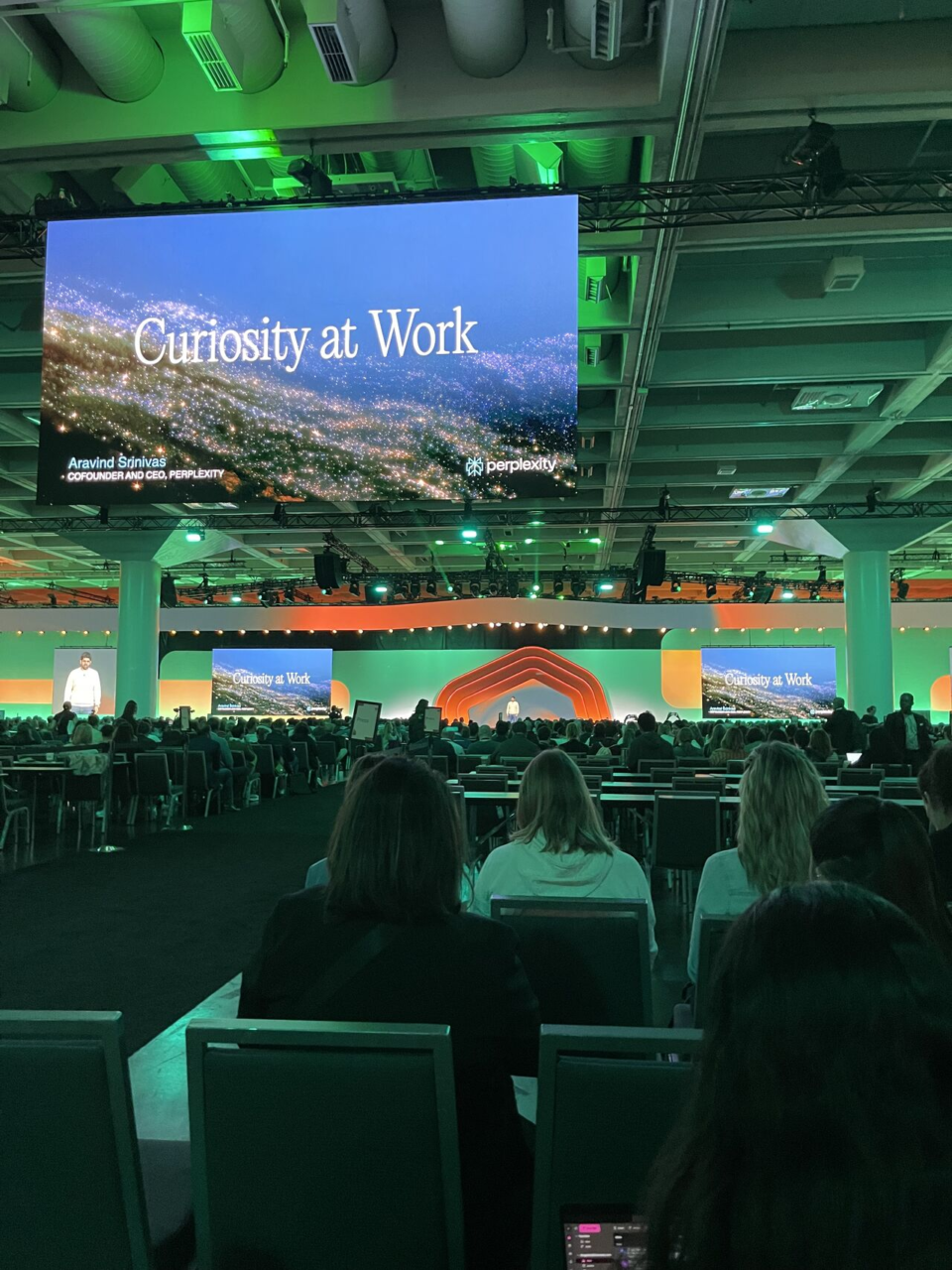 A large audience facing a stage screen that reads Curiosity at Work