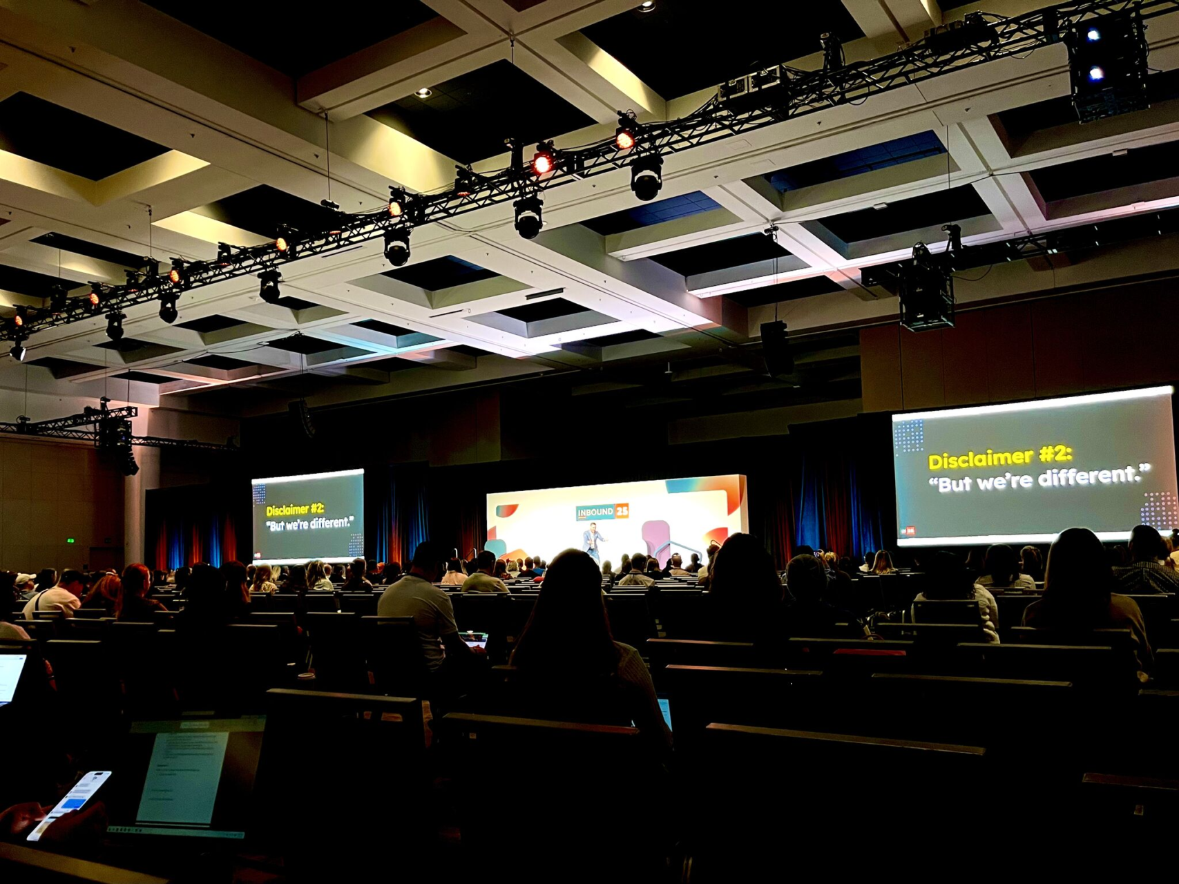 A ballroom audience watching a session, with slides that read Disclaimer #2: But we're different