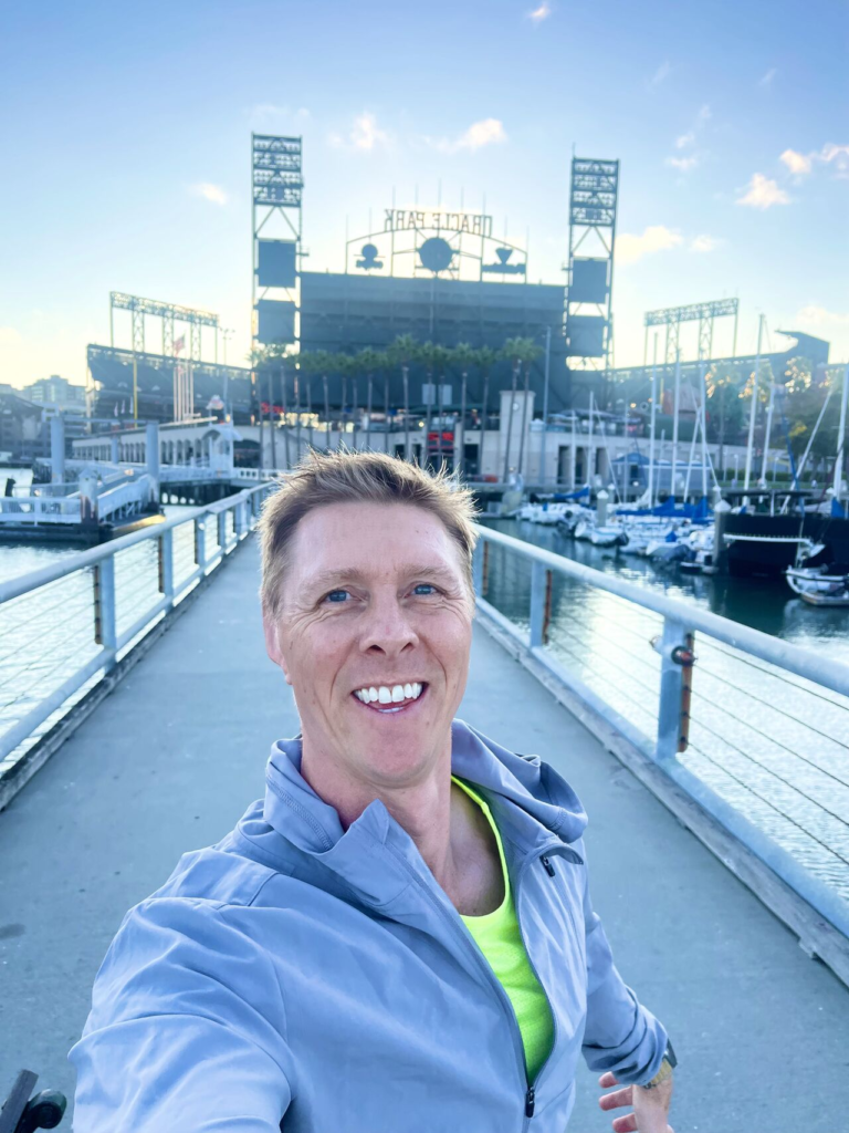 A smiling man takes a selfie on a waterfront pier with a ballpark behind him