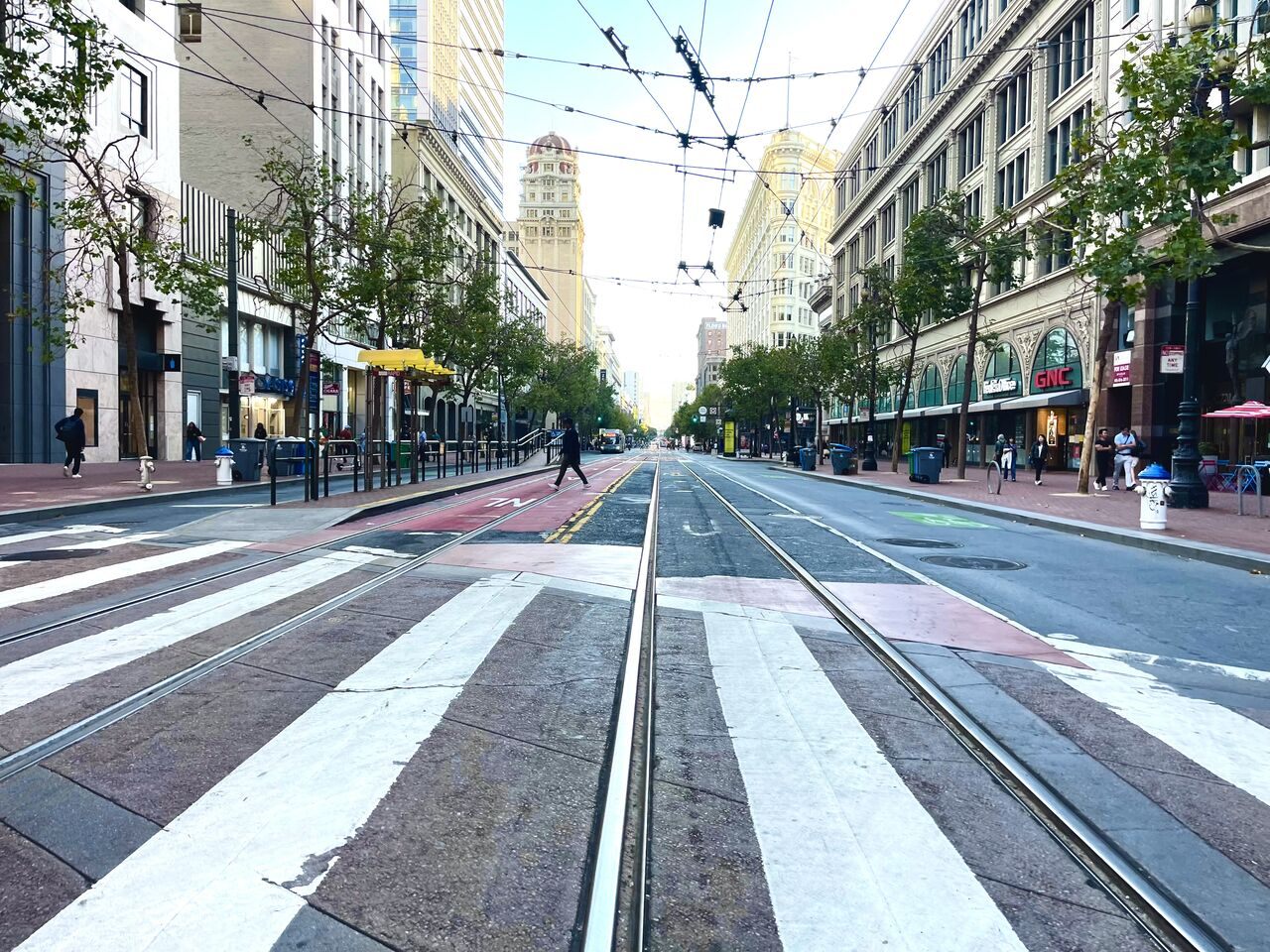 A wide city street with crosswalk stripes and streetcar tracks, lined with shops
